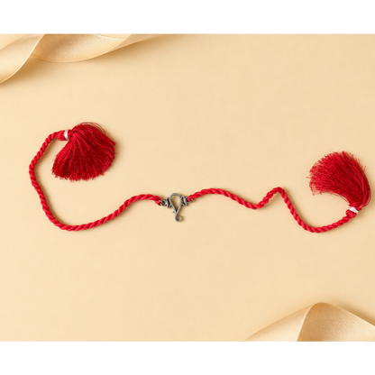 A red braided cord ornament with two red tassels and a small silver clasp, arranged on a warm beige background with soft cream ribbon curls in the corners.