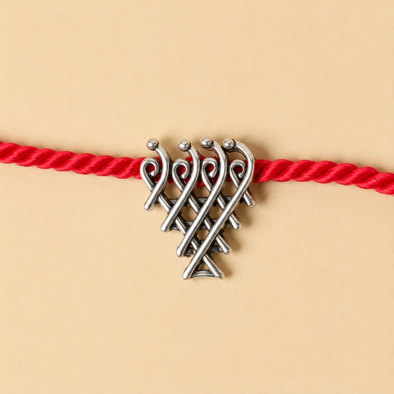 A silver-toned metal knot charm with small bead accents, threaded onto a bright red braided cord, shown against a warm beige background.
